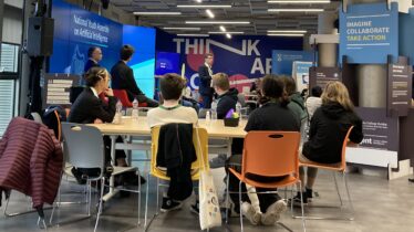 A group of young students sitting around a table listening to people talk about AI